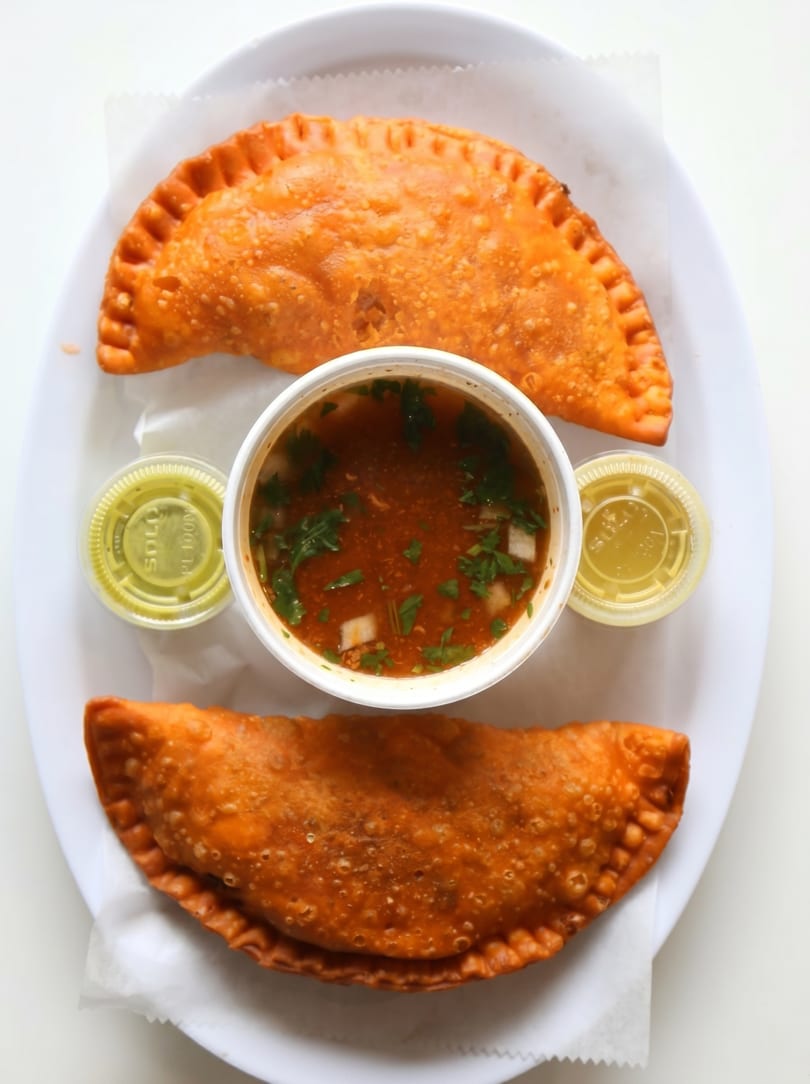 Two golden-brown empanadas flank a cup of red broth with cilantro and two small green sauce containers.